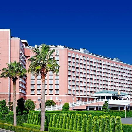 Готель Grand Nikko Tokyo Bay Maihama Urayasu Екстер'єр фото The image shows a large, multi-story hotel building featuring a light pink facade. The architecture appears modern, with numerous windows arranged in a grid pattern. In the foreground, there are neatly trimmed shrubs and palm trees, adding a tropical