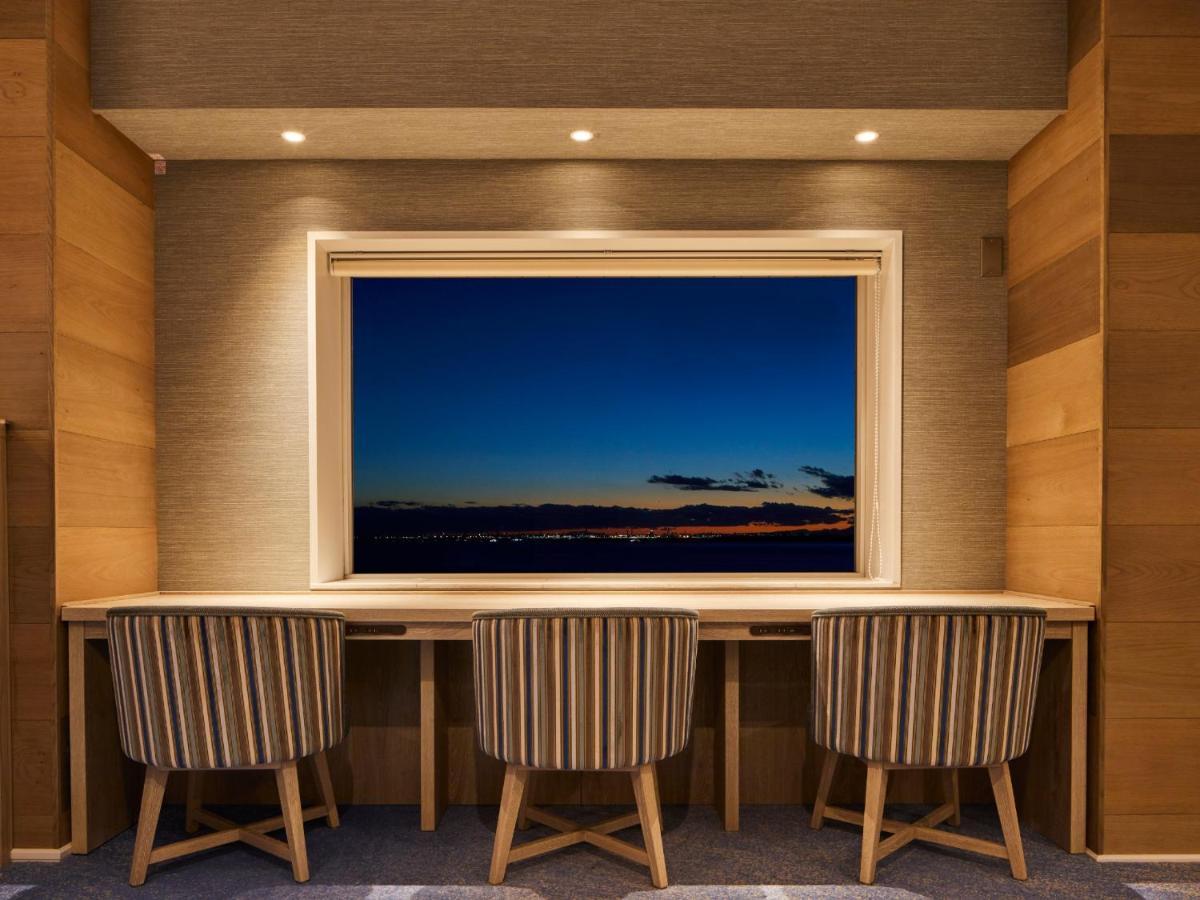 Готель Grand Nikko Tokyo Bay Maihama Urayasu Екстер'єр фото The photo depicts a cozy workspace or study area featuring a long, minimalist desk with three chairs. The wall behind the desk is made of wood, creating a warm atmosphere. At the center, there is a large window that showcases a beautiful evening view