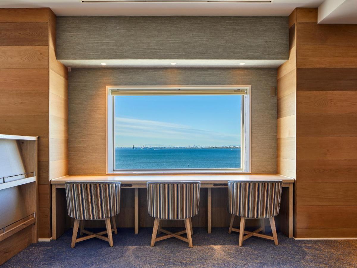 Готель Grand Nikko Tokyo Bay Maihama Urayasu Екстер'єр фото The photo shows a modern workspace with three striped chairs positioned at a long, white desk. The desk faces a large window with a view of a tranquil body of water and a distant skyline. The walls around the workspace are made of wood paneling, crea