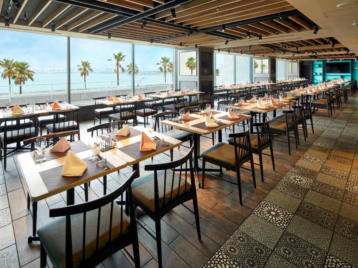Готель Grand Nikko Tokyo Bay Maihama Urayasu Екстер'єр фото The photo shows a modern restaurant interior featuring rows of neatly arranged tables and chairs. The large windows provide a scenic view of palm trees and what appears to be a body of water in the background, suggesting a coastal location. Each tabl