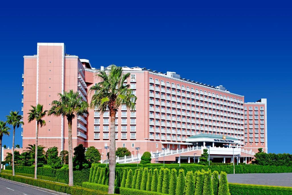 Готель Grand Nikko Tokyo Bay Maihama Urayasu Екстер'єр фото The image shows a large, multi-story hotel building featuring a light pink facade. The architecture appears modern, with numerous windows arranged in a grid pattern. In the foreground, there are neatly trimmed shrubs and palm trees, adding a tropical